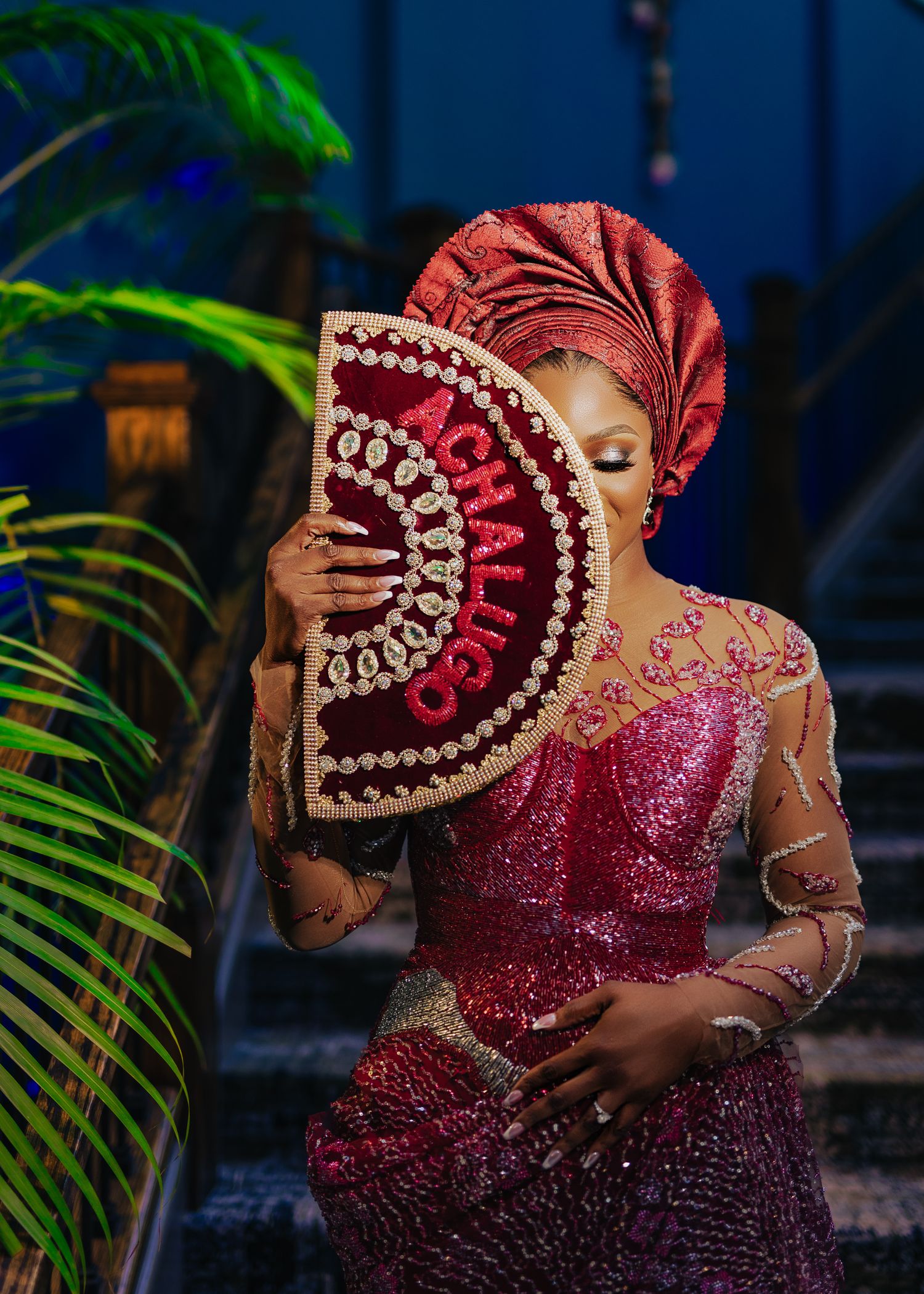 Elegant figure in a sparkling red gown and matching headwrap poses with a decorative fan against a blue background.