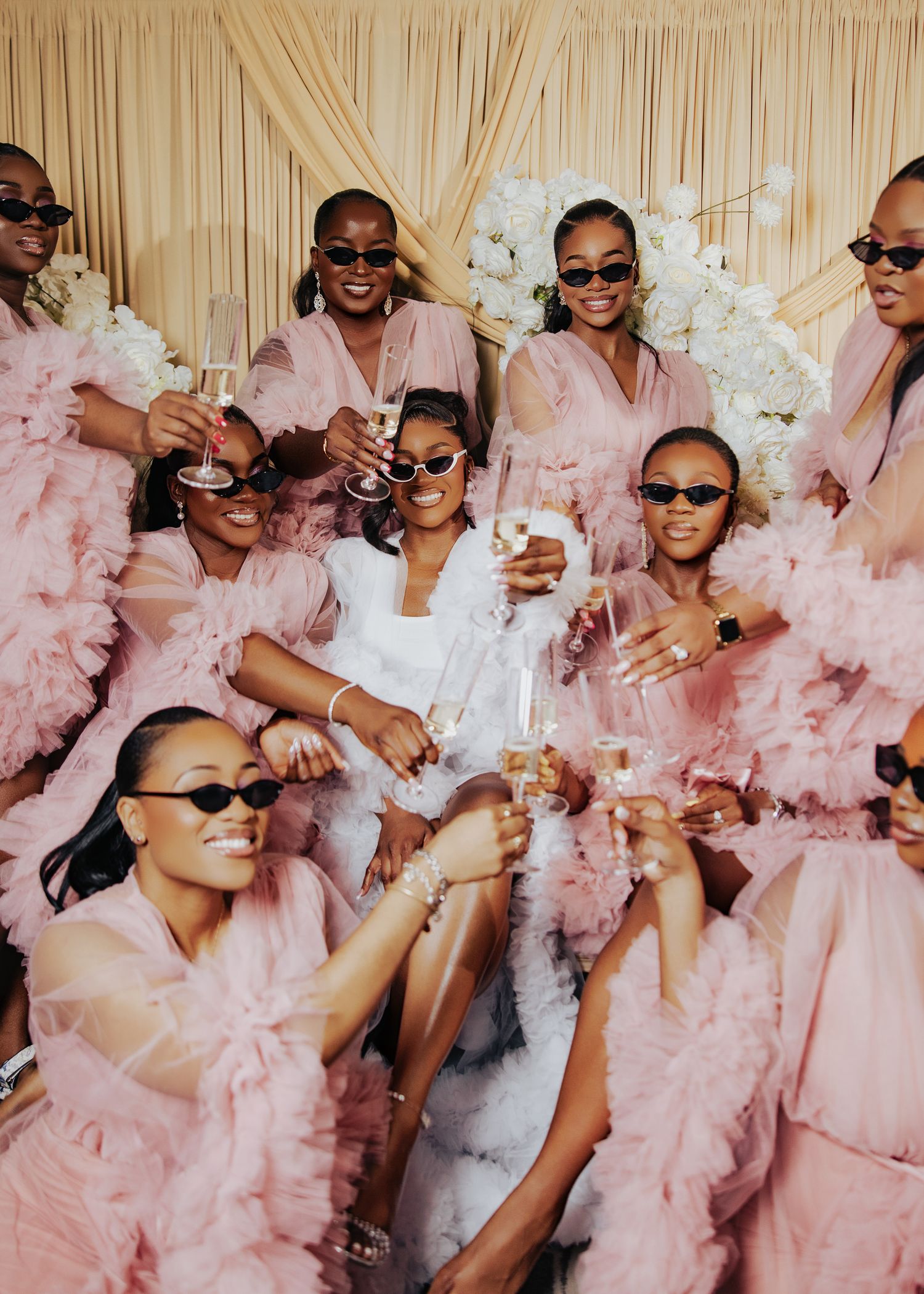 Bridal party wearing pink dresses and sunglasses poses playfully with champagne glasses.