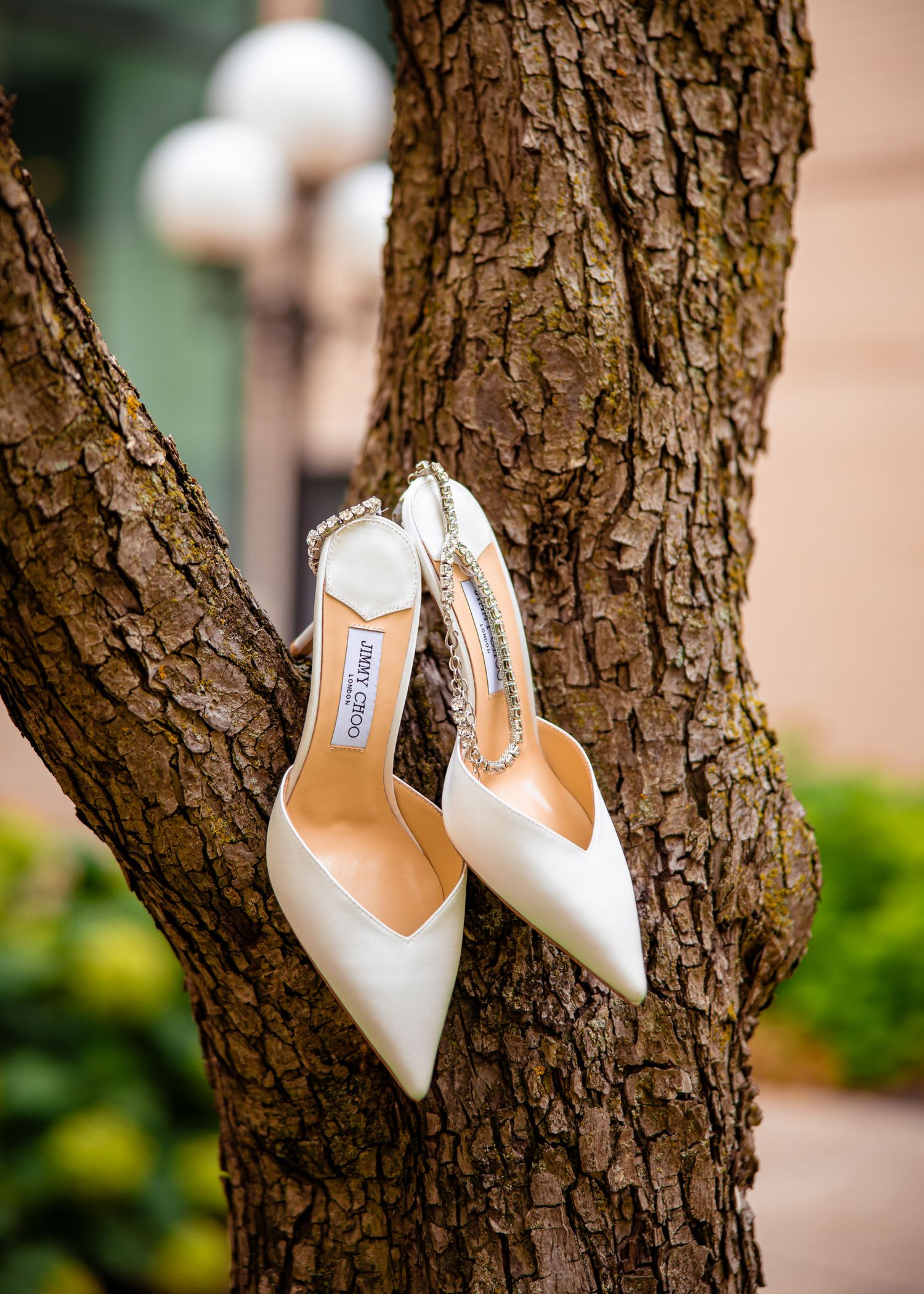 White satin bridal heels elegantly displayed on a textured tree trunk in a garden setting.