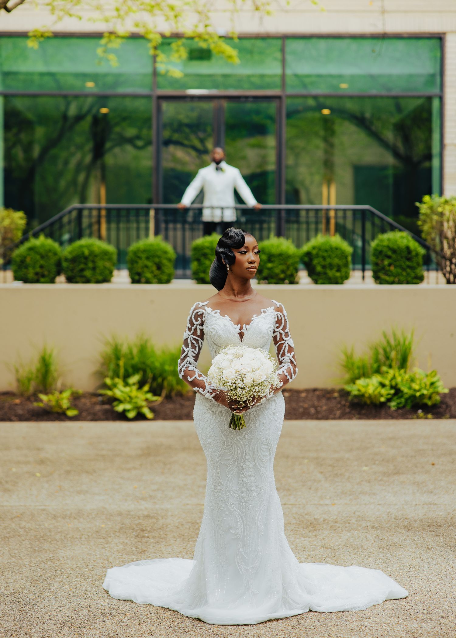 Bride in elegant lace-sleeved mermaid wedding gown holding white bouquet against modern building backdrop.