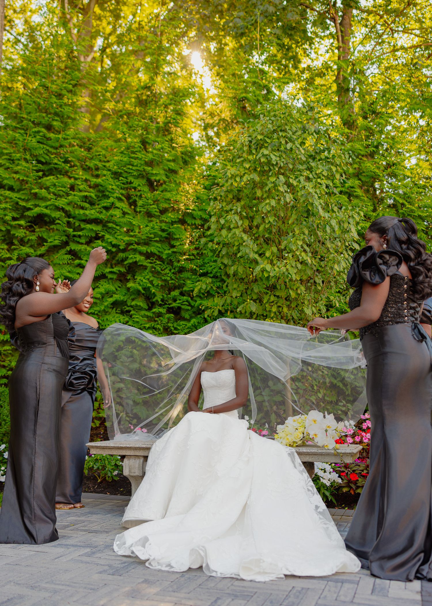Wedding party in dark dresses helps arrange bride's veil in outdoor garden setting.