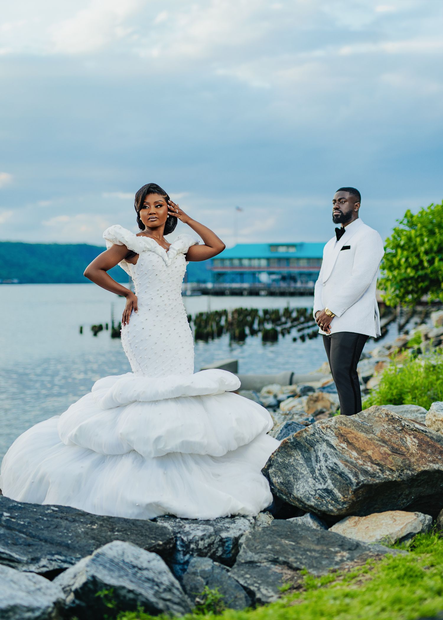 A couple poses for romantic wedding photos on rocky coastal terrain with a pier and ocean in the background.