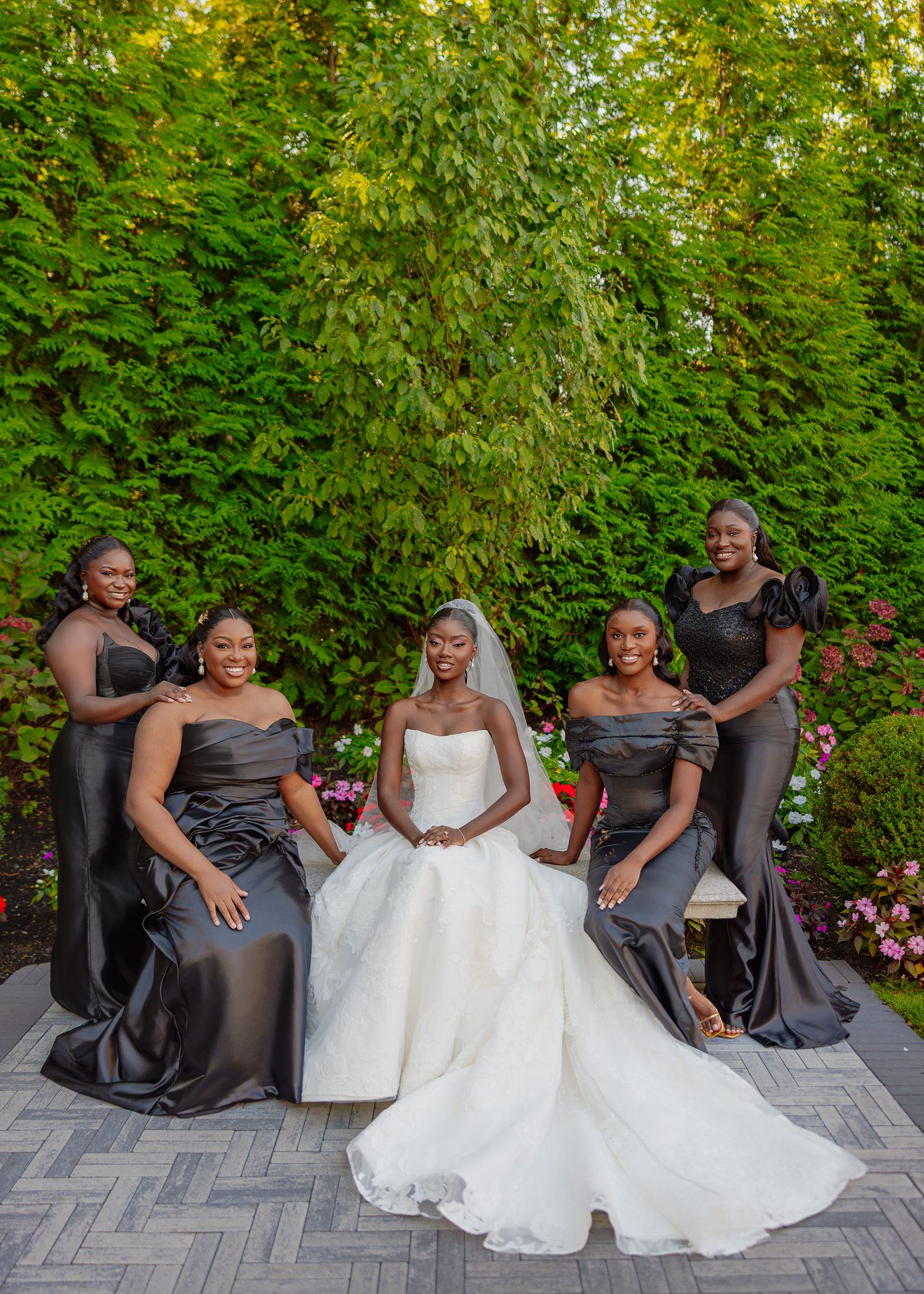 Wedding party in dark gray bridesmaid dresses and white bridal gown sit together on stone steps in garden setting.