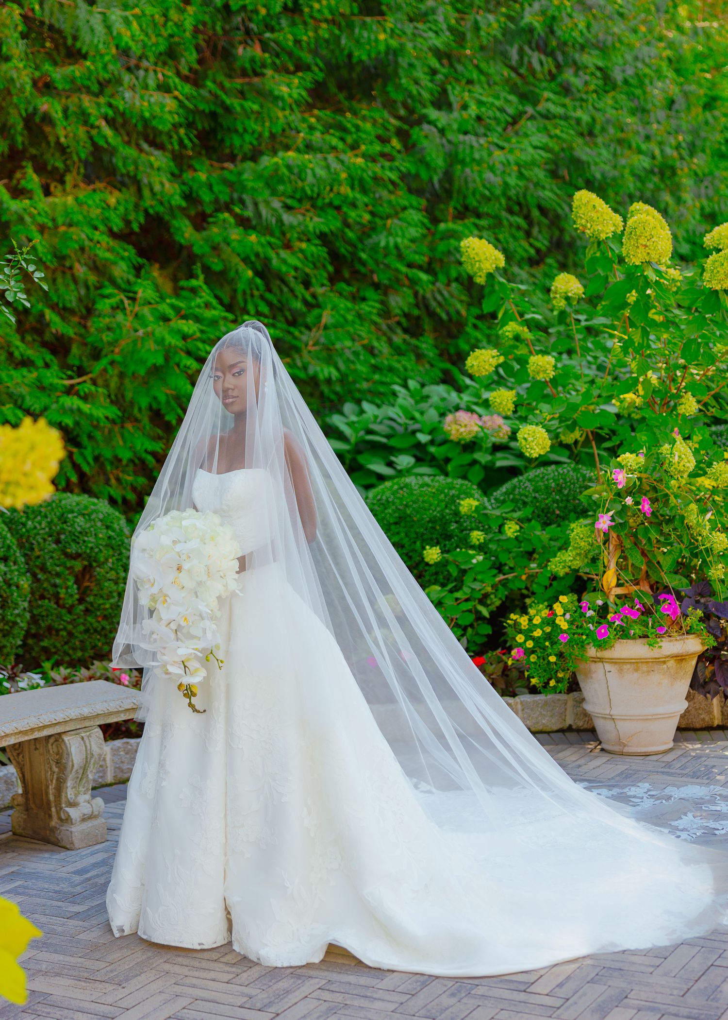 Bride in classic white gown with cathedral veil stands gracefully on stone pathway surrounded by vibrant greenery.