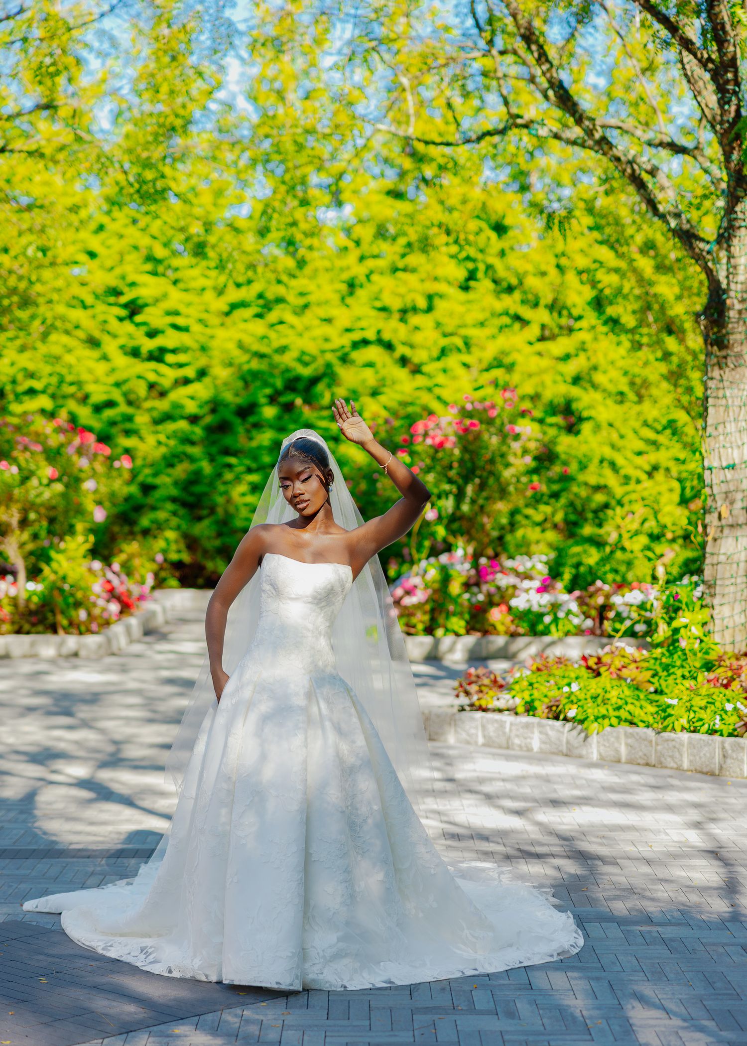 Joyful bride twirls in voluminous ballgown on garden pathway with yellow flowers and trees in background.