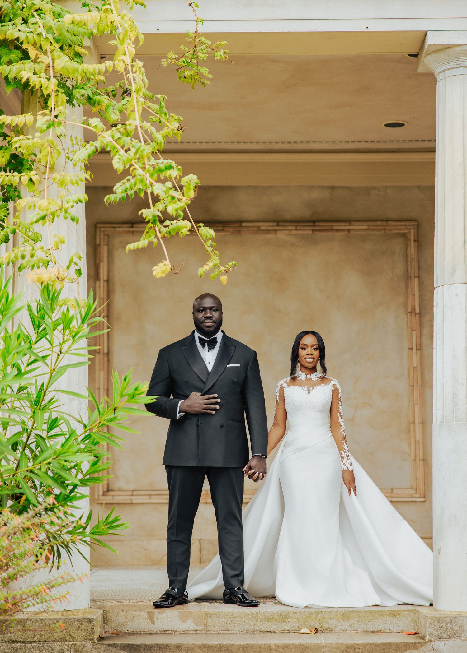 Elegant couple in formal wedding attire pose under a classic white columned portico.