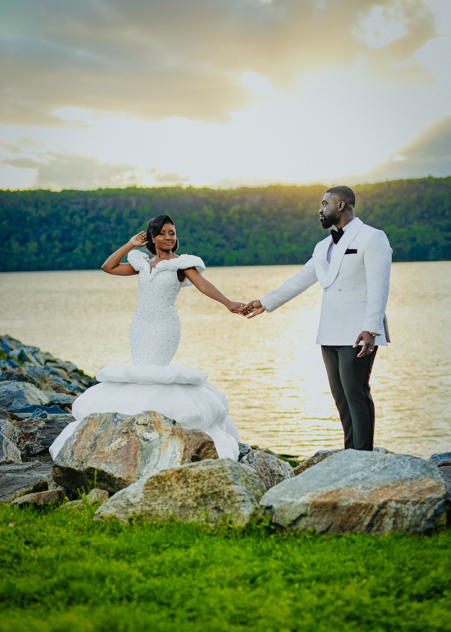 A romantic sunset wedding photo of a couple holding hands while standing on rocky terrain by a lake.