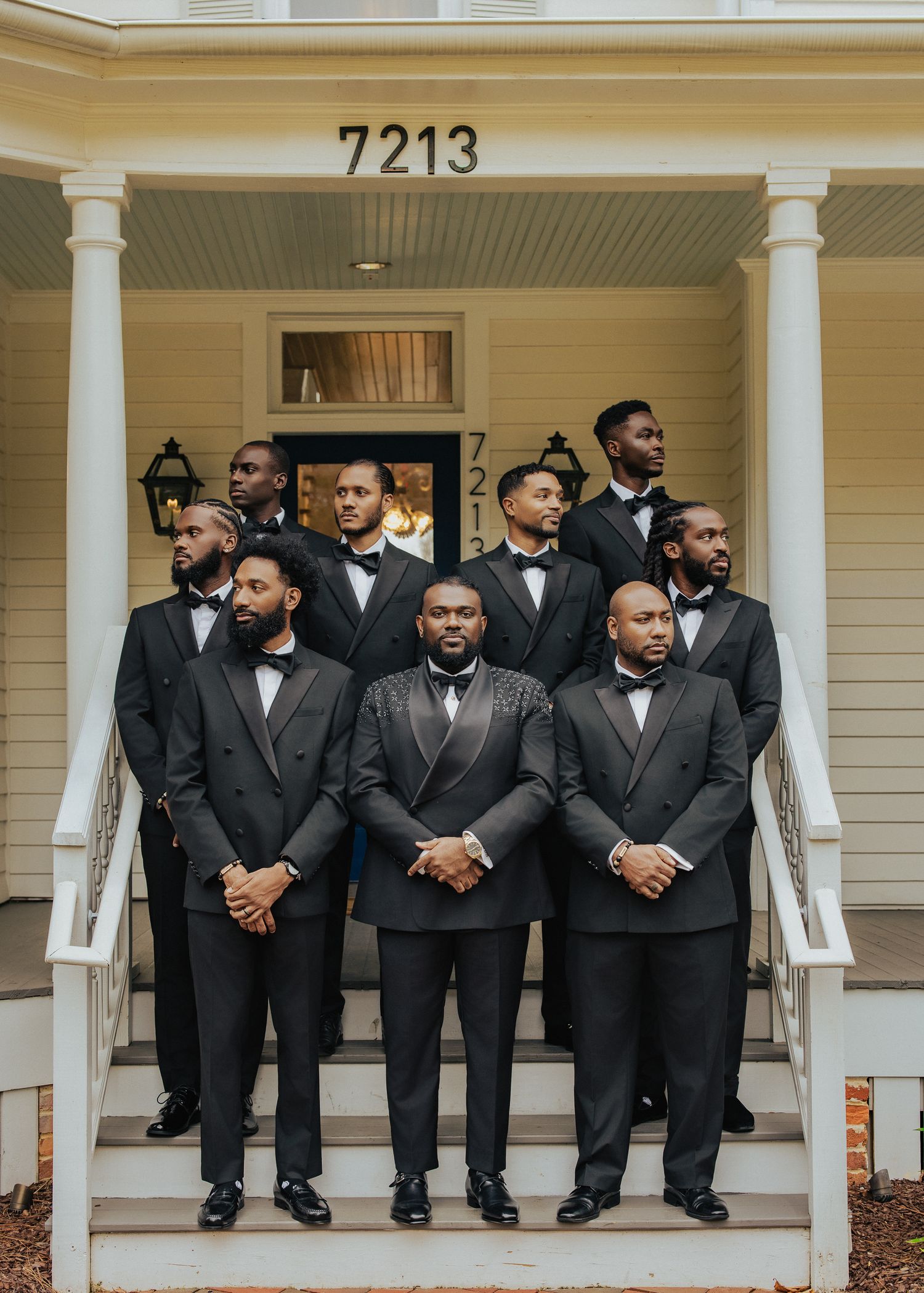 Groomsmen in black tuxedos and bow ties stand together on a white porch with columns at house number 7213.