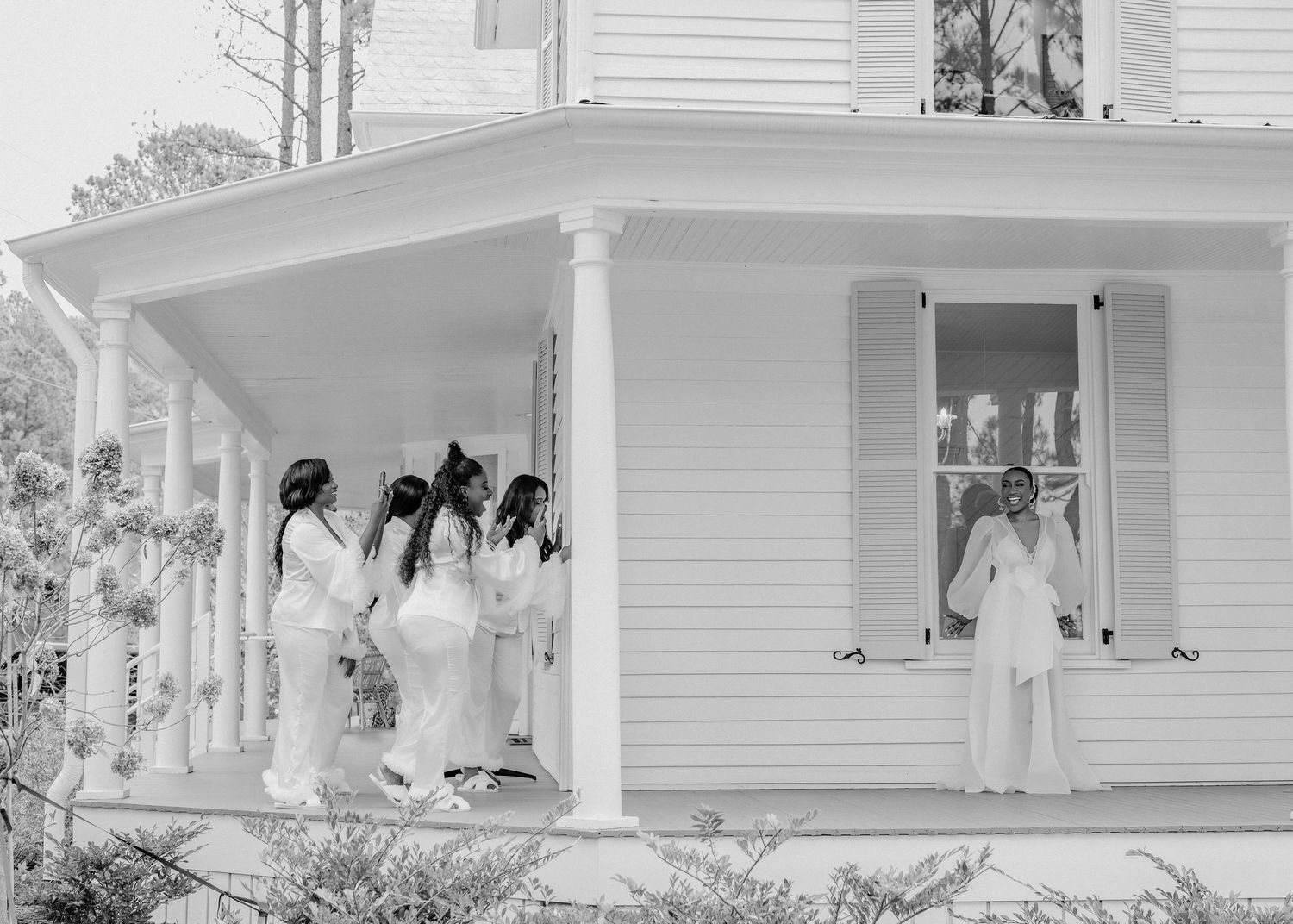 A group in white dresses gathers on the porch of a classic white clapboard house with columns and shutters.