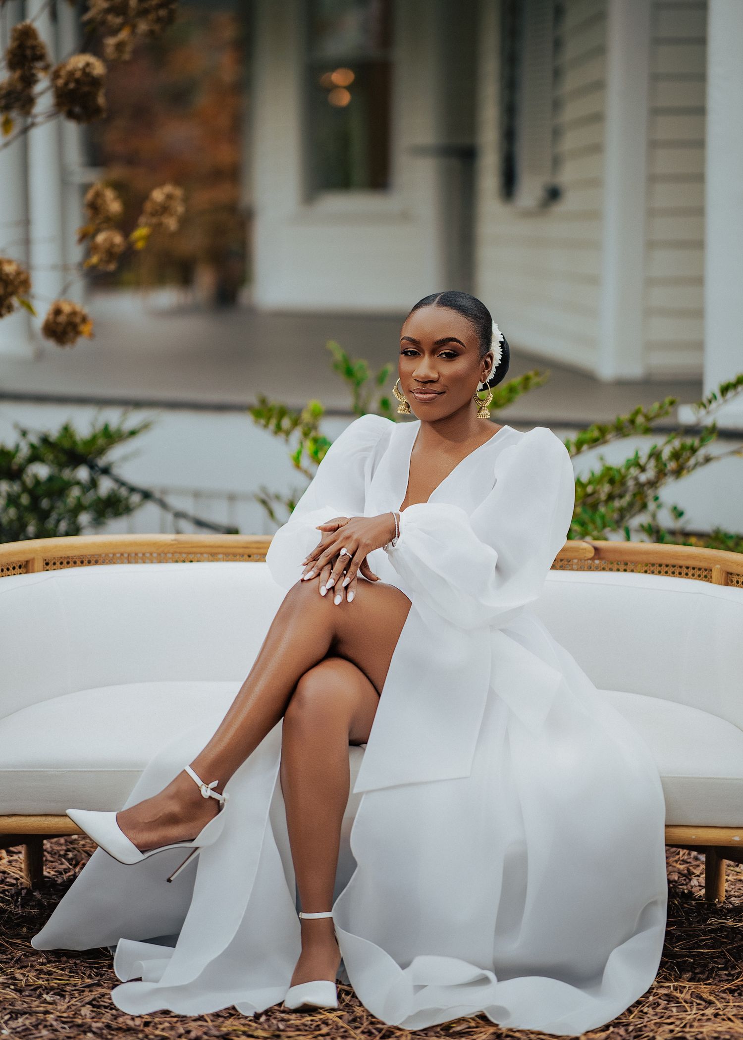 Elegant bride in flowing white dress lounges on outdoor patio furniture during autumn wedding photoshoot.