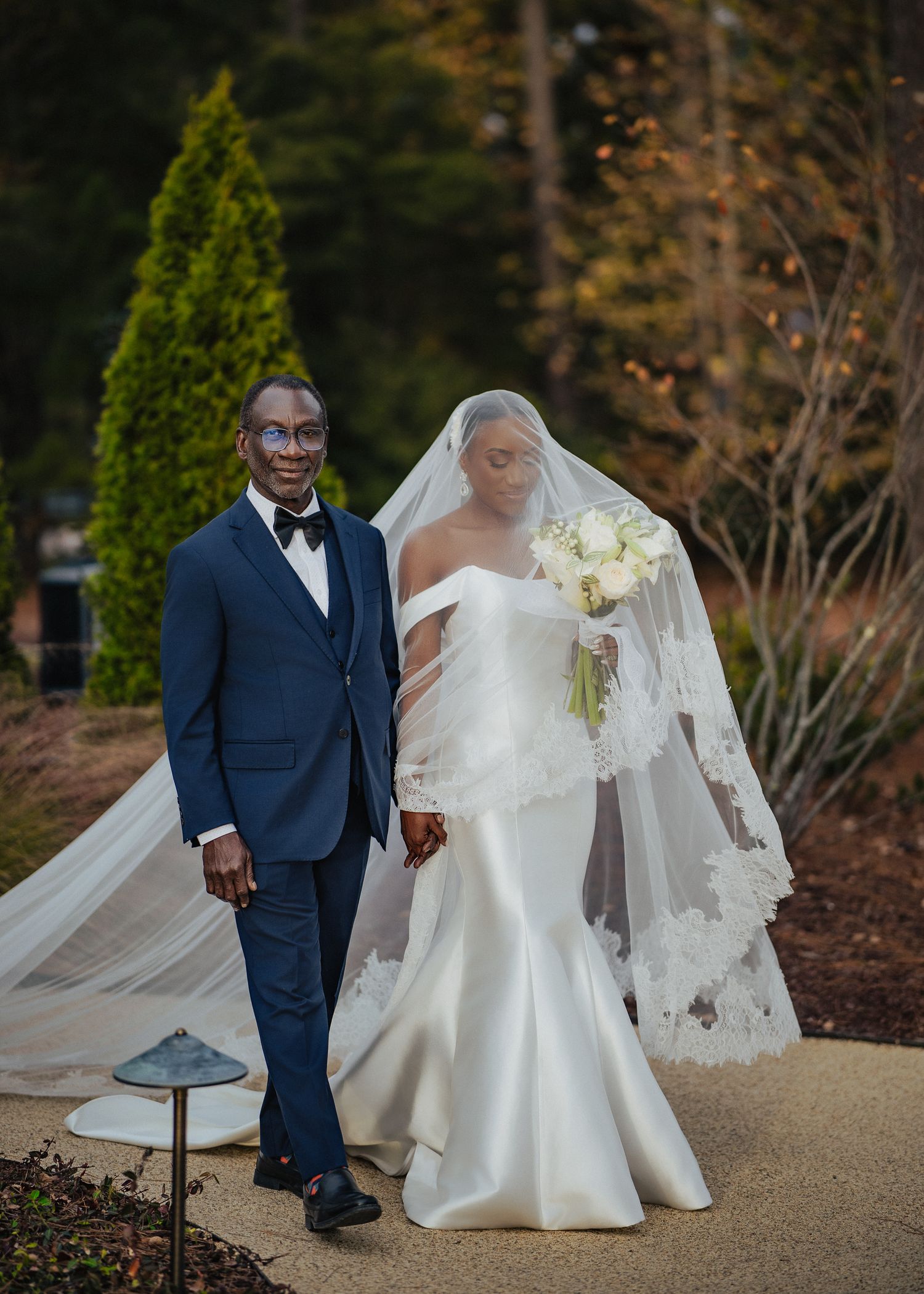 A bride in an elegant white gown and groom in a navy tuxedo pose together on a garden path during their wedding.
