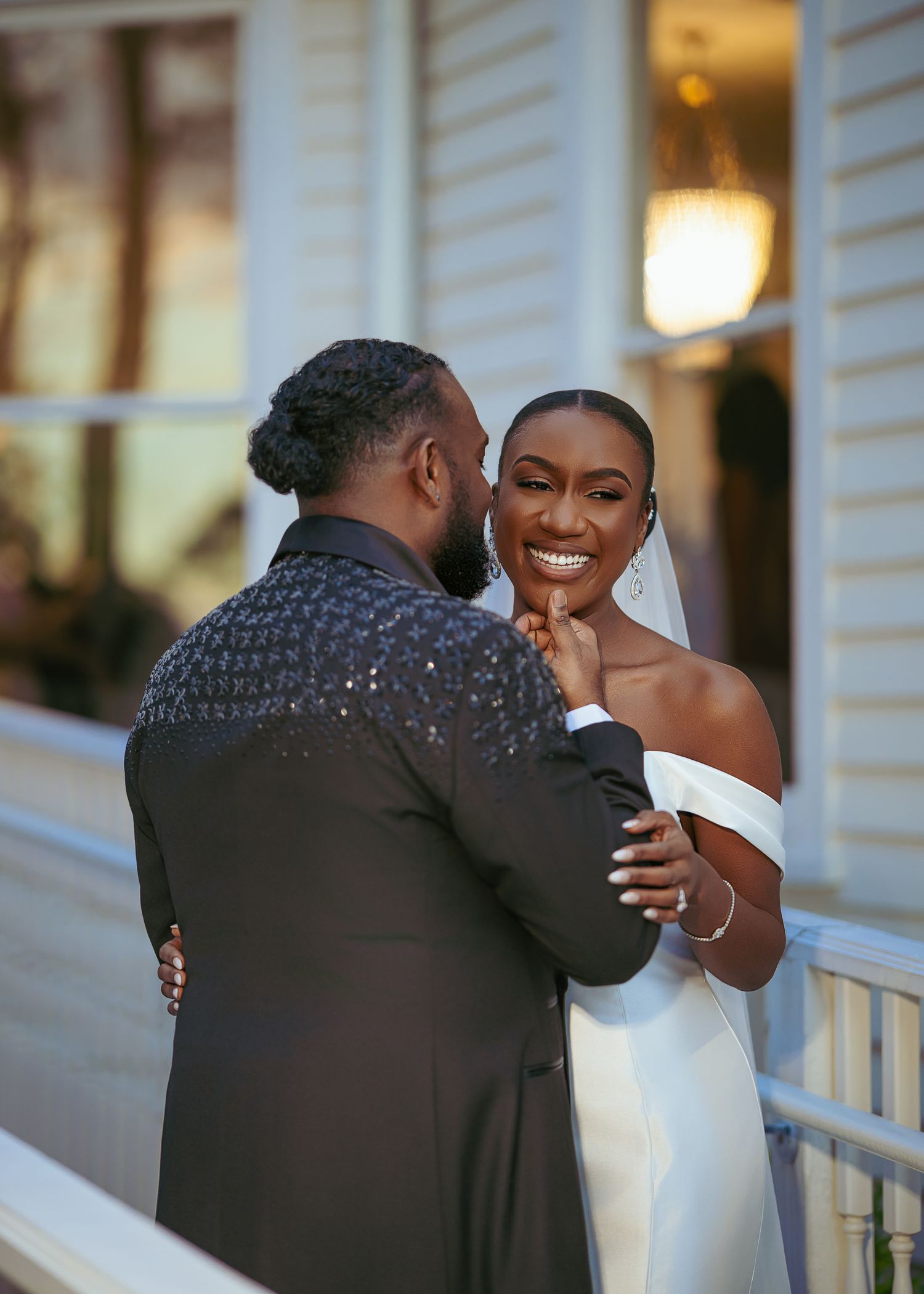 A romantic moment between a couple in wedding attire sharing laughter and embraces on a white porch at sunset.