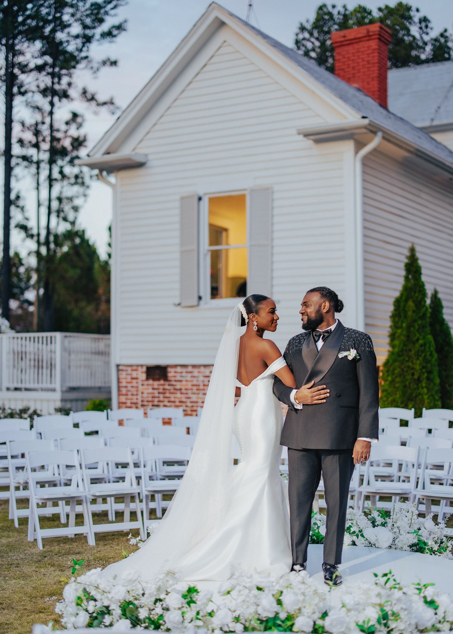 A joyful wedding couple shares an intimate moment outside a white chapel with rows of white chairs and floral decor.