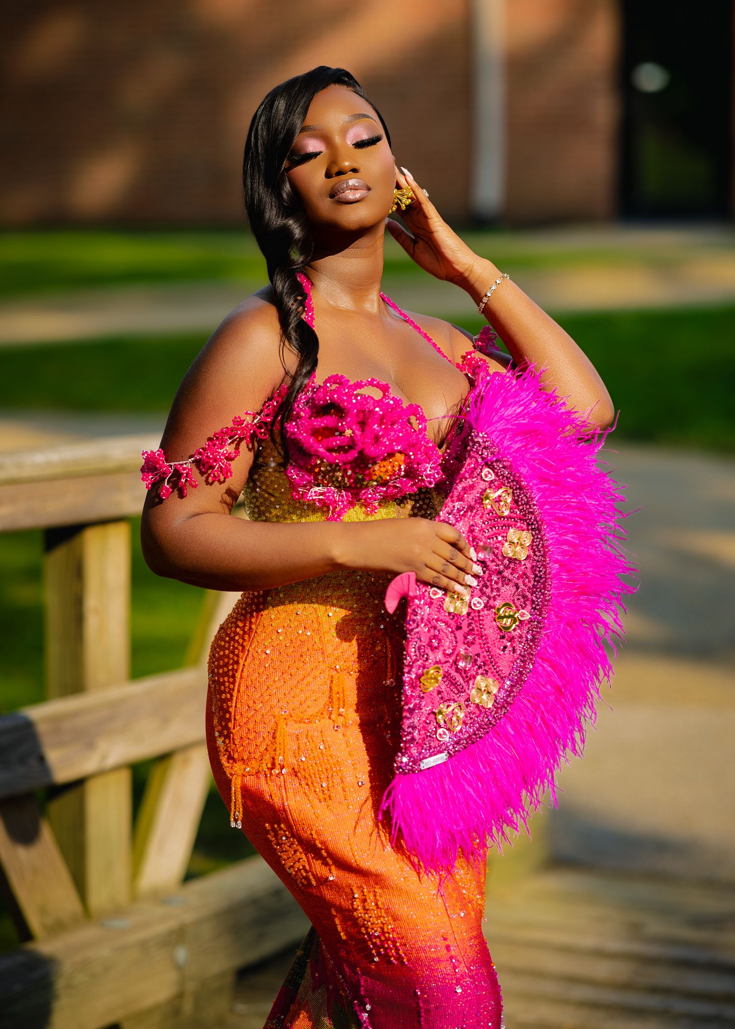 Glamorous model in bright pink and orange sequined gown with feather fan poses at golden hour on wooden bridge.