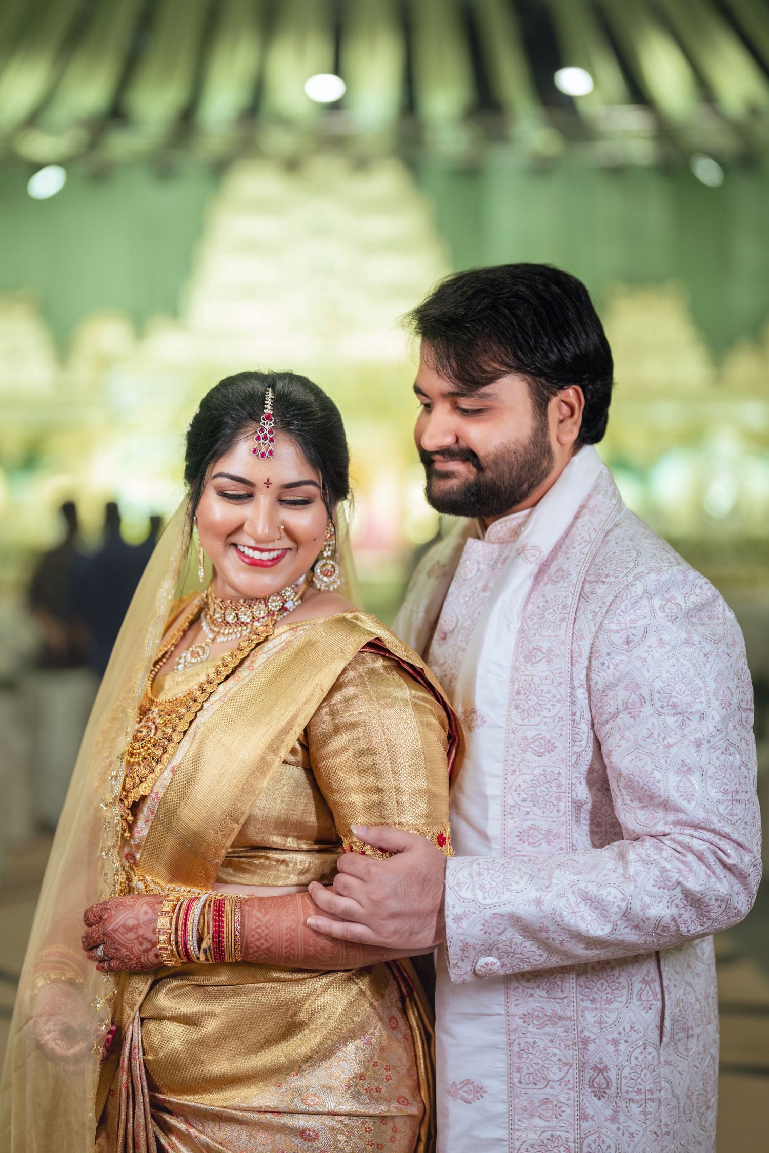 South Indian wedding couple in traditional gold and cream attire pose under decorative green lighting.