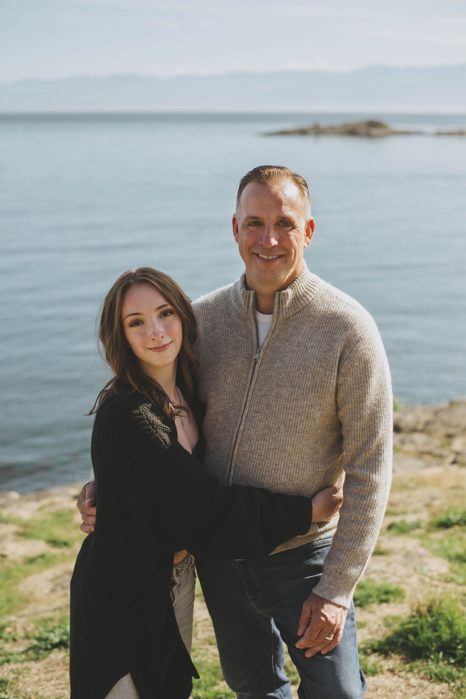 Father and daughter standing close together by the ocean, smiling on a sunny day.