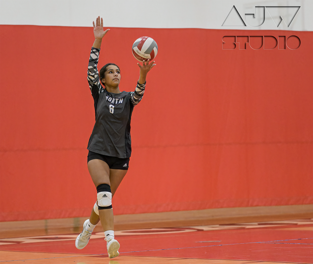 Volleyball player in black uniform jumps to serve against a bright red gymnasium wall.