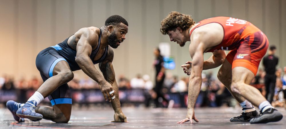 Two wrestlers in blue and orange singlets face off in starting position on the competition mat.