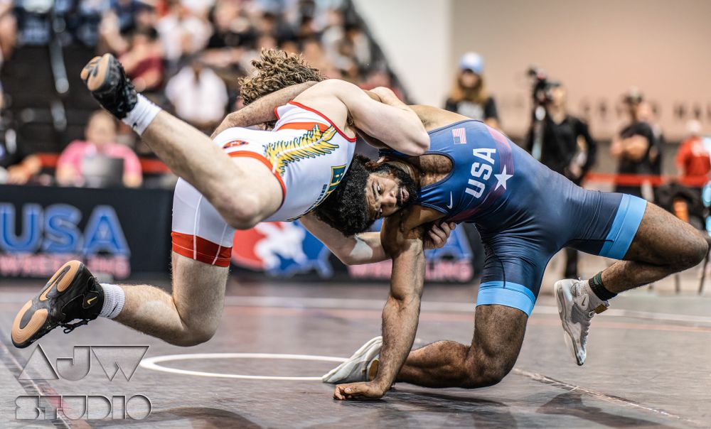 Wrestling match action shows athletes grappling in blue and red uniforms on competition mat.