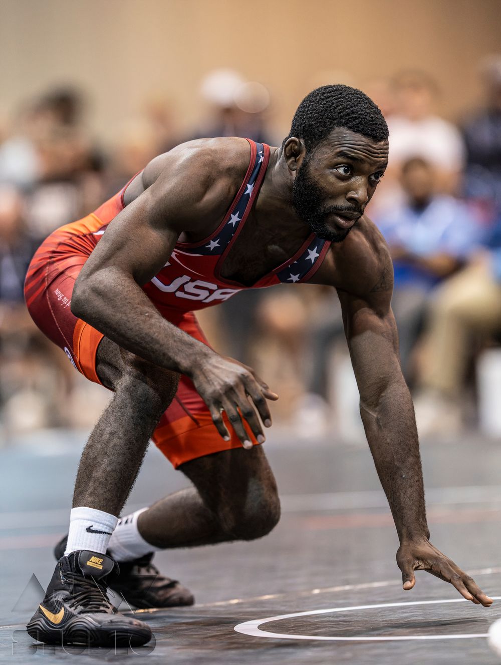 Wrestler in red and blue USA uniform crouches in starting position on wrestling mat during competition.
