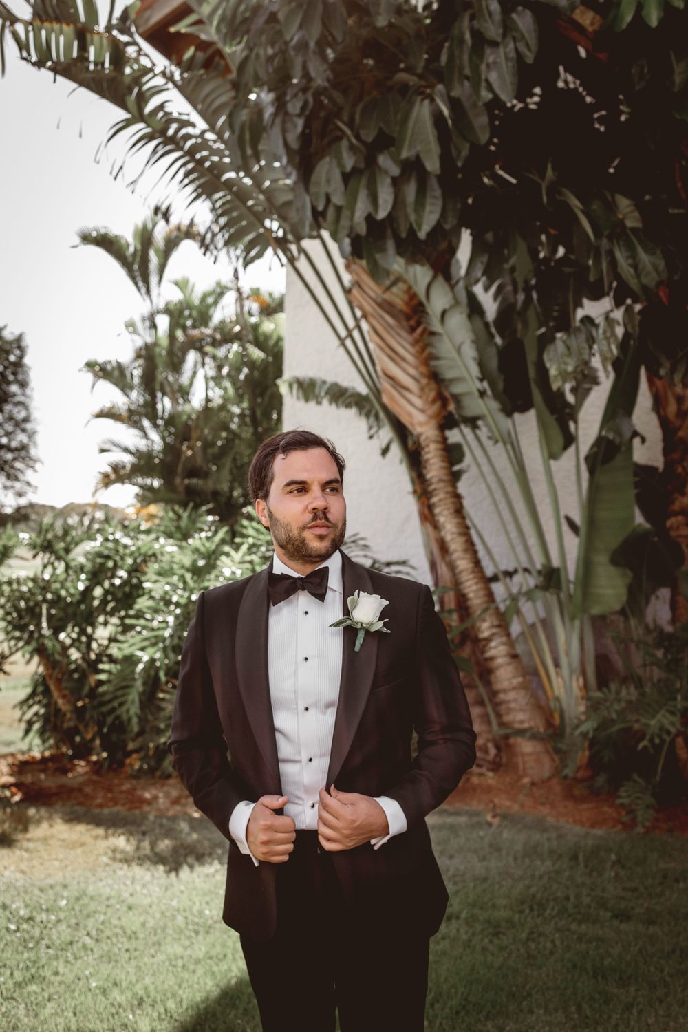 Groom in black tuxedo poses against tropical palm trees in outdoor wedding setting.