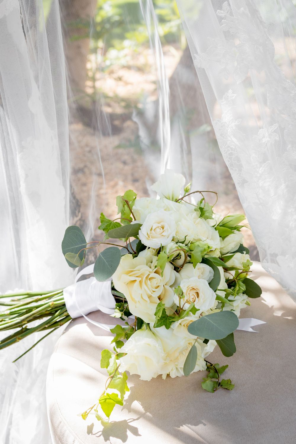 White rose and eucalyptus wedding bouquet resting on sheer white fabric in natural sunlight.