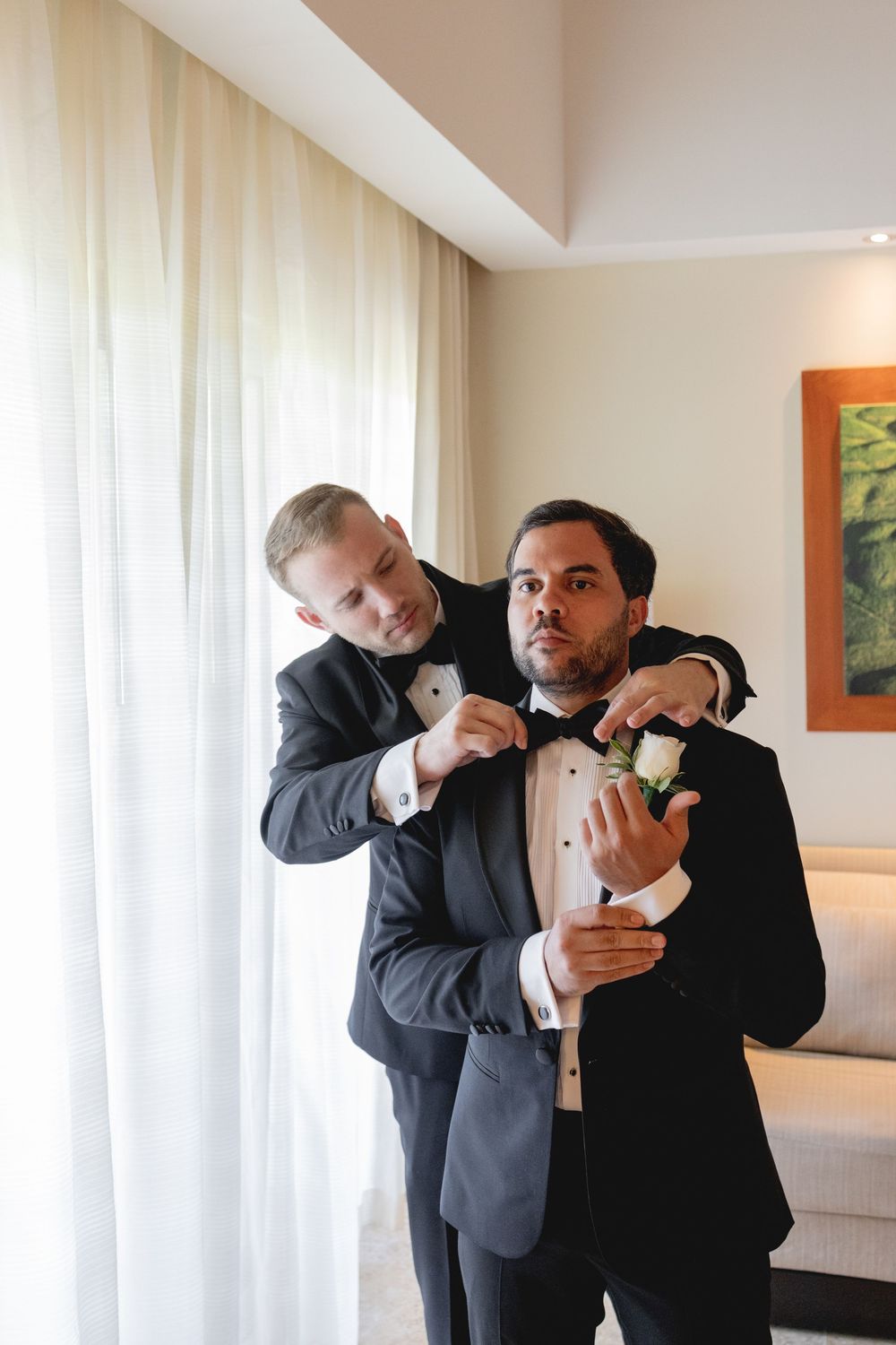 Two men in tuxedos playfully adjust each other's bow ties in a bright hotel room.