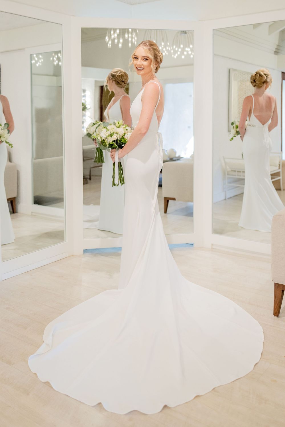 A stunning white wedding dress with a flowing train displayed in an elegant white room with mirrors.