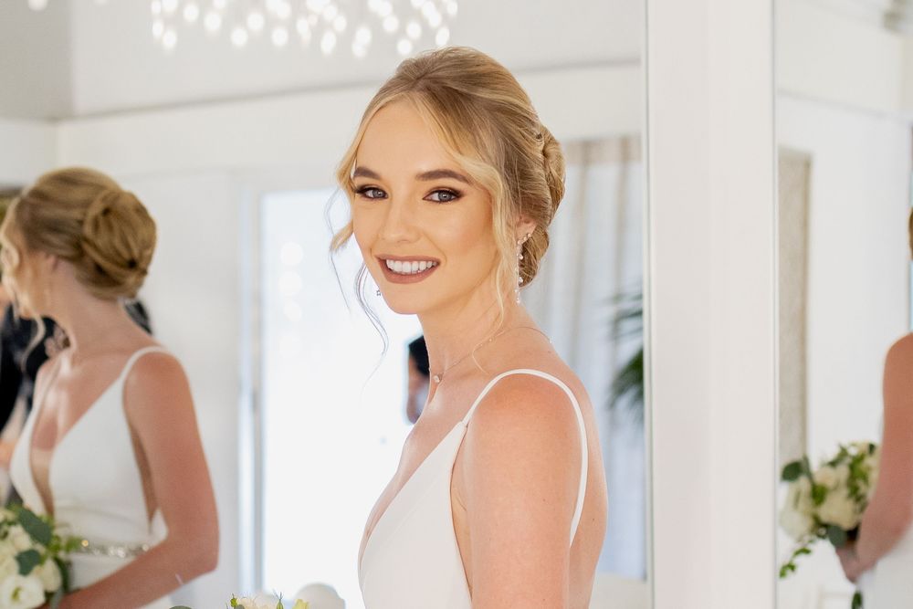 A radiant bride wearing a white dress smiles brightly in a sunlit room with a mirror reflection in the background.