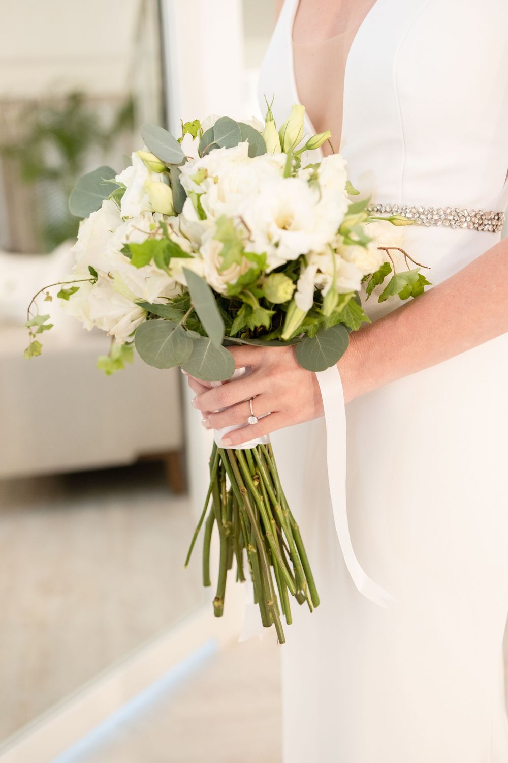 Elegant white bridal bouquet with roses, eucalyptus leaves and ribbon being held against a white wedding dress.