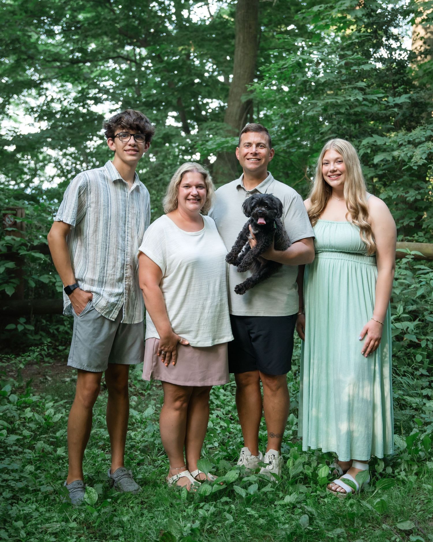 A family group sits together on a white blanket outdoors surrounded by green foliage.