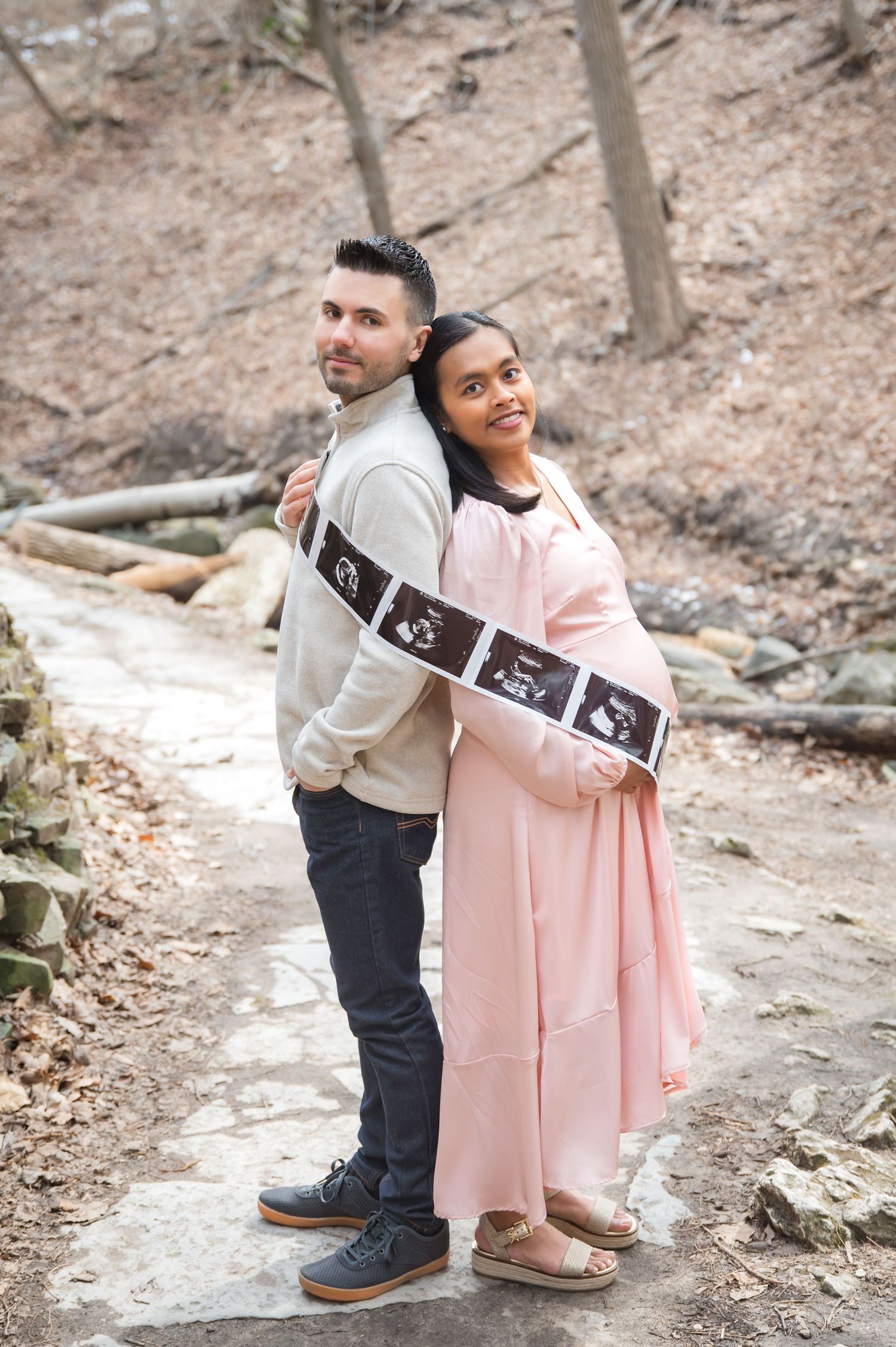 A couple stands back-to-back outdoors holding ultrasound photos in a pregnancy announcement photoshoot.