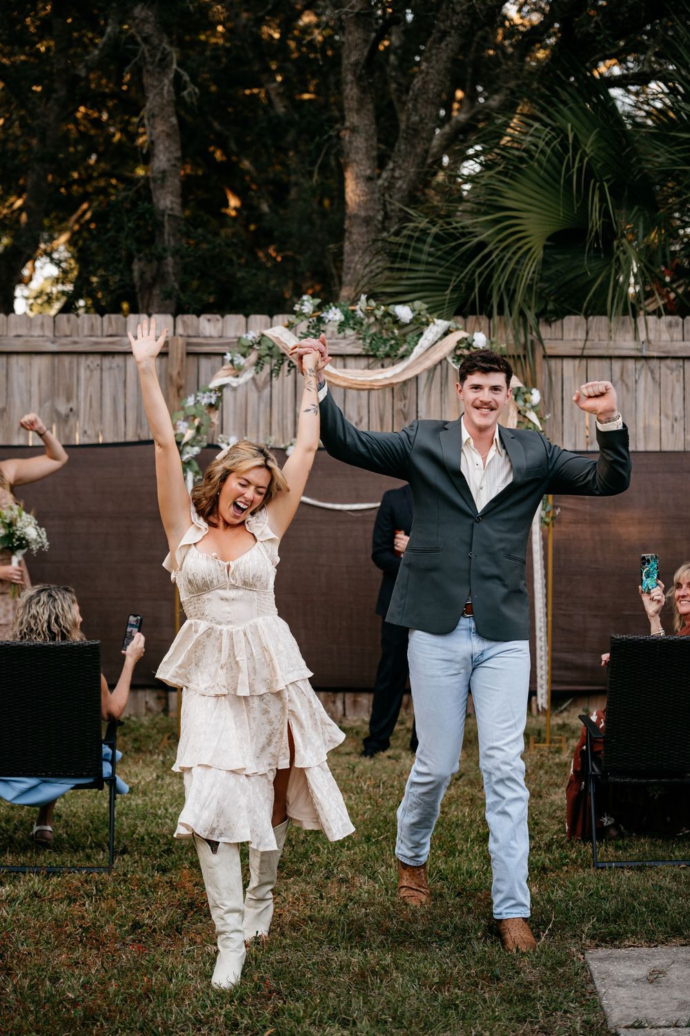 Playful couple dancing and celebrating in a backyard garden setting with wooden fence and palm trees.