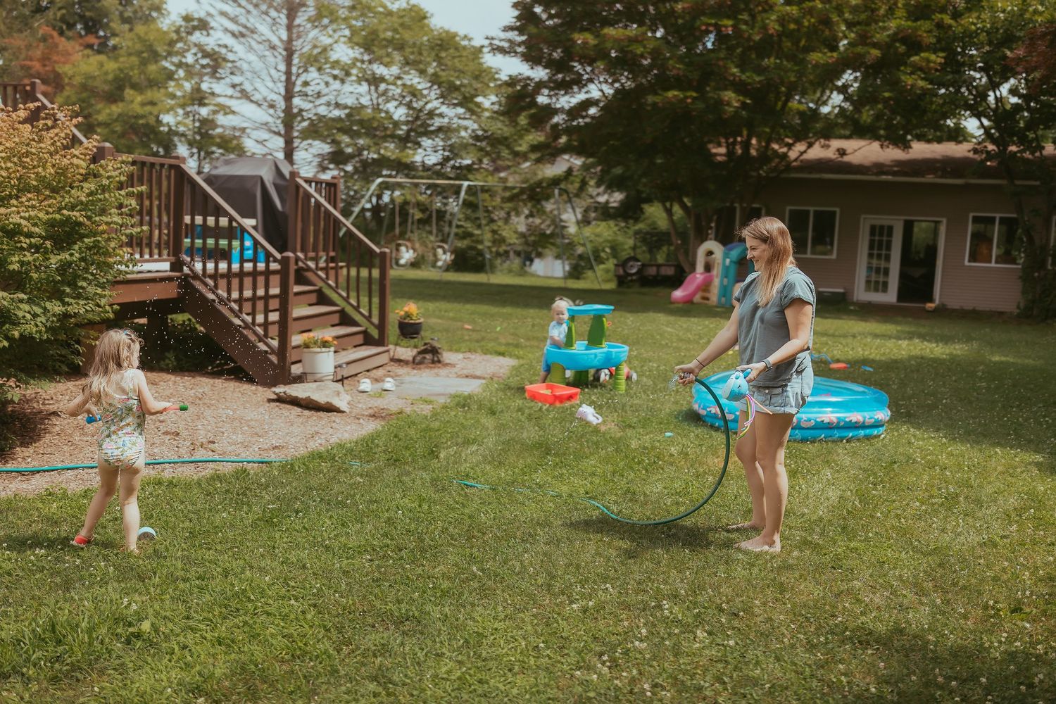 People playing with water toys and sprinklers in a sunny backyard with stairs and green grass on a summer day.