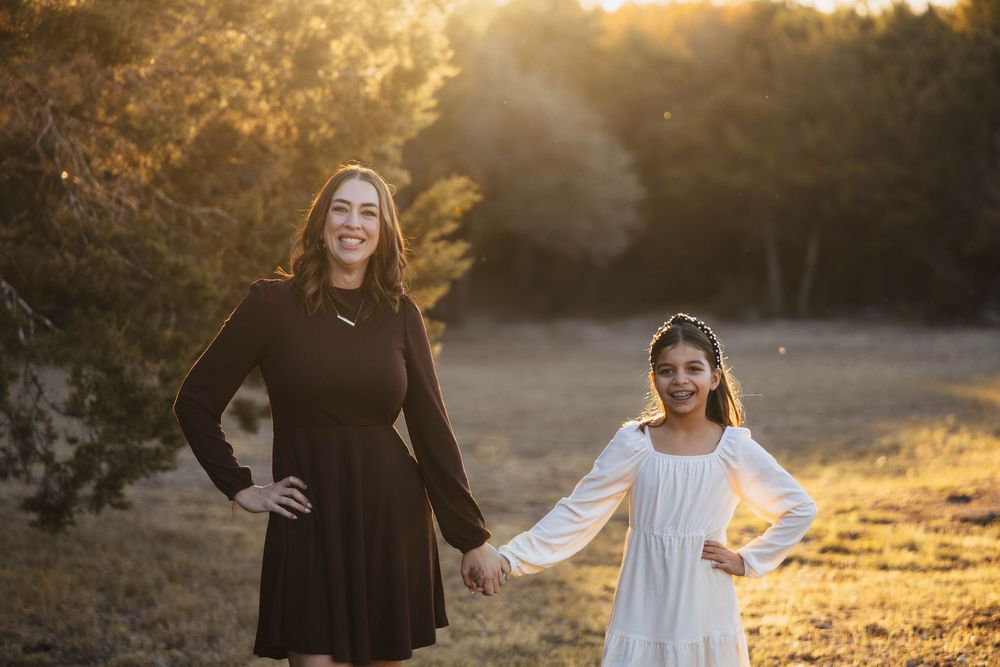 Two people in dresses hold hands while standing in a golden sunlit field at sunset.