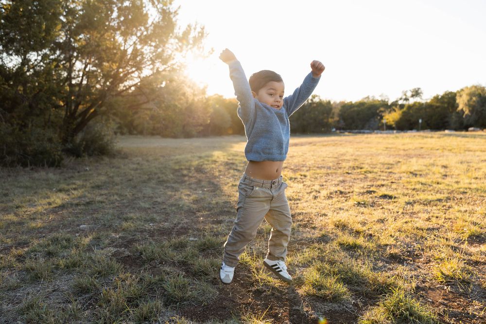 Silhouetted figure jumps joyfully in a sunlit meadow at golden hour with trees in background.