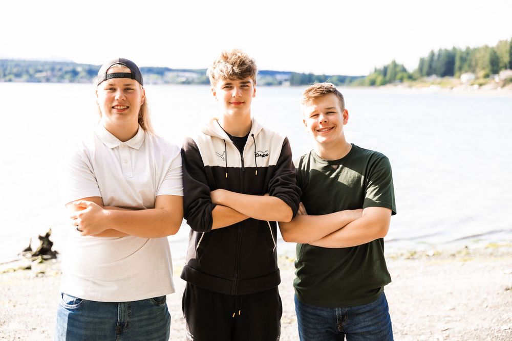 Three people stand with crossed arms and smiling on a sunny beach with a lake and trees in the background.