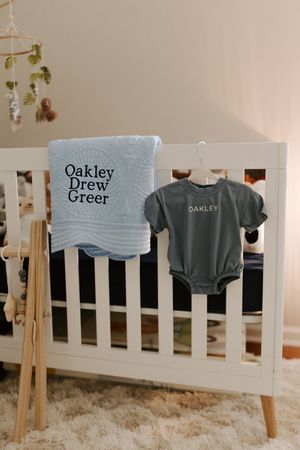 Gray t-shirts hanging on white crib railing displaying personalized text.