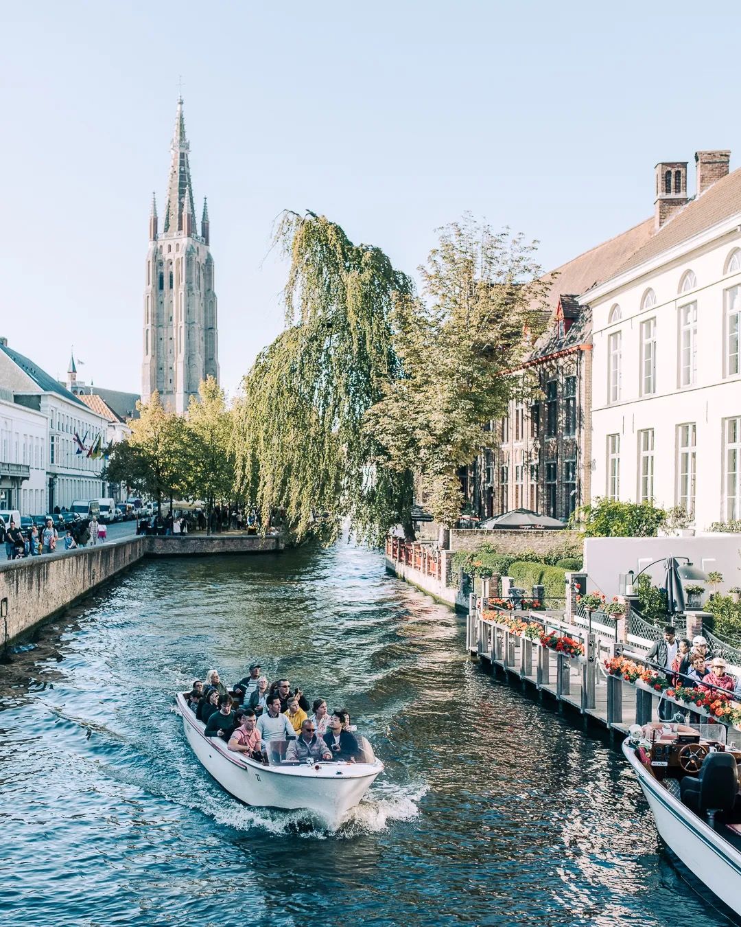 Boat of tourists on a canal with historic buildings and a tall church spire.