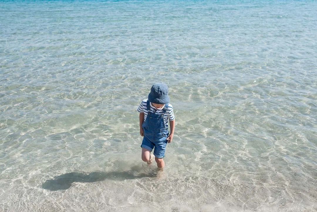 A young child standing in crystal-clear shallow seawater under bright summer light.