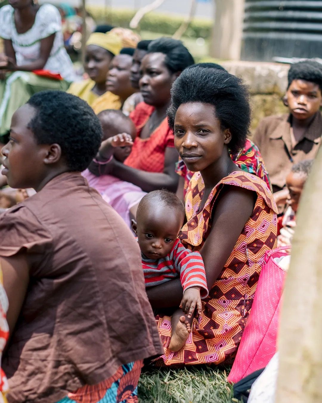 Women and children sit together outdoors, watching an event and sharing a relaxed community moment.