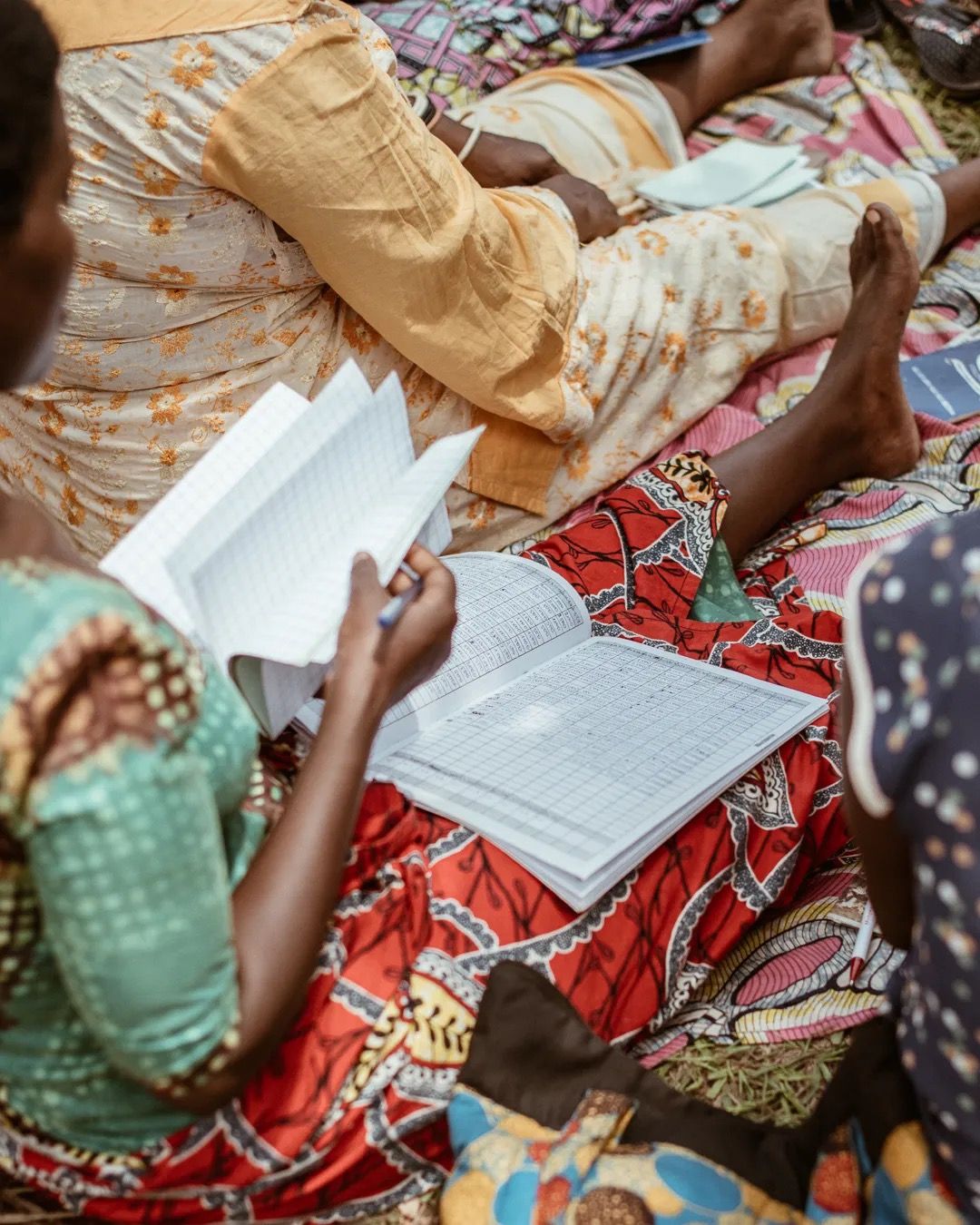 Women sit together reading and writing in notebooks, showing community learning and education.