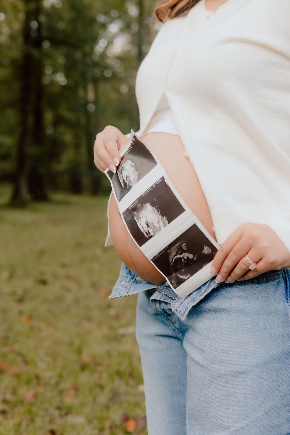 Person in blue jeans and white top holding ultrasound photos outdoors in a natural setting.