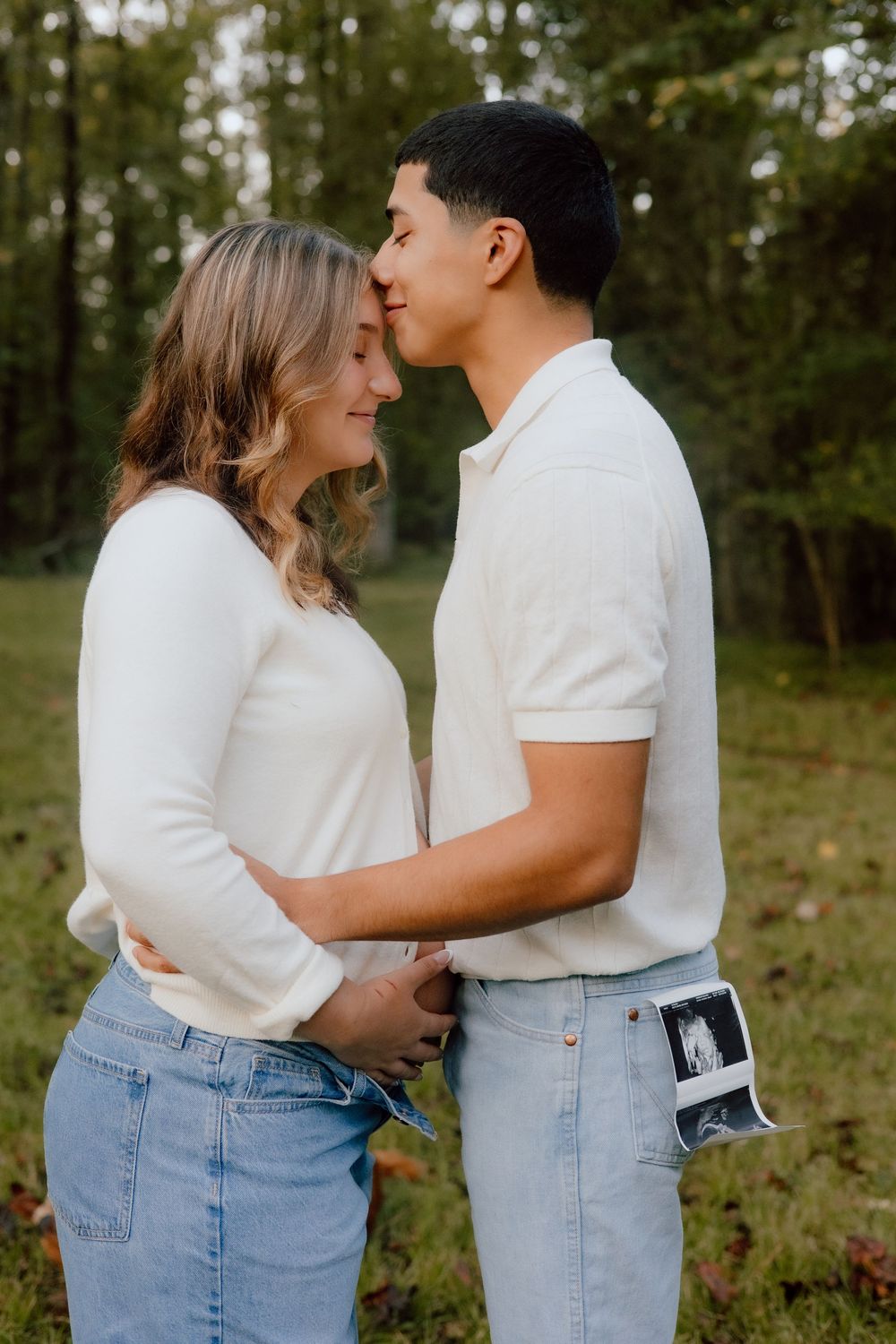 A romantic couple in matching white tops and jeans share an intimate moment outdoors in a wooded setting.