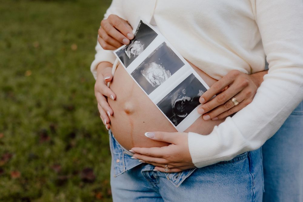 Hands holding ultrasound photos while wearing a cozy white sweater and blue jeans during a pregnancy announcement photoshoot.