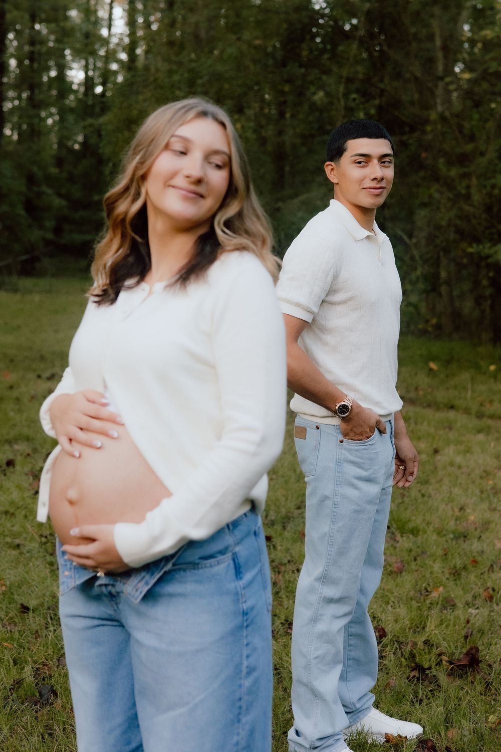 A couple in white tops and jeans embraces tenderly in a natural outdoor setting with trees in the background.