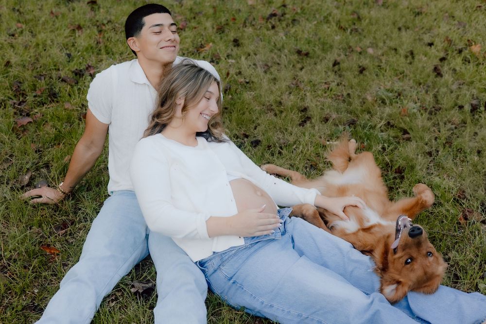 A couple in white and denim relaxes on grass with their playful brown dog on a sunny day.