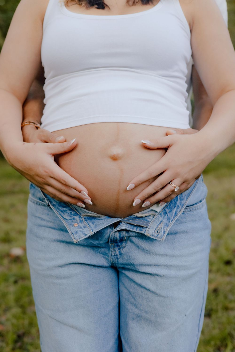 Close up of a pregnant belly with hands gently cradling the bump while wearing jeans and a white top.