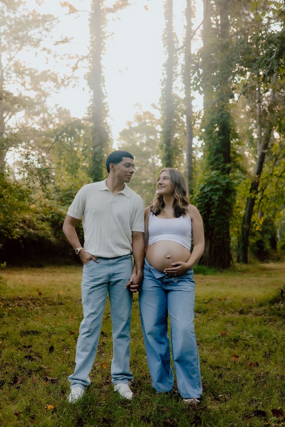 A couple in casual attire stands together in a sunlit wooded area during an outdoor photo session.