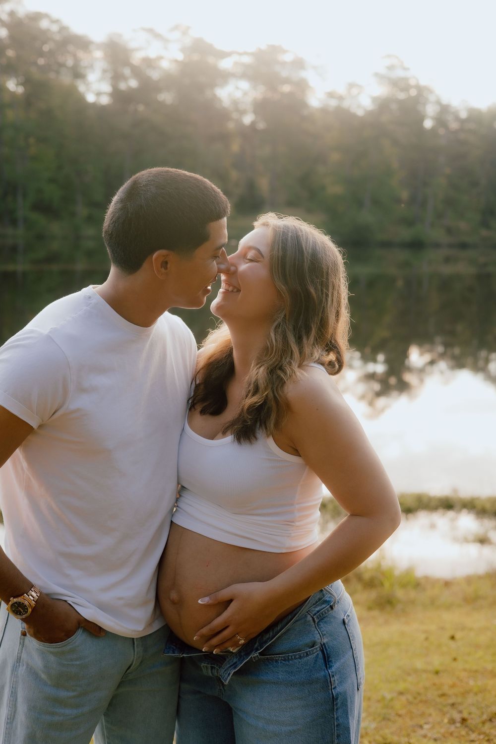 A couple in casual white tops and jeans embraces lovingly by a serene lake during golden hour.